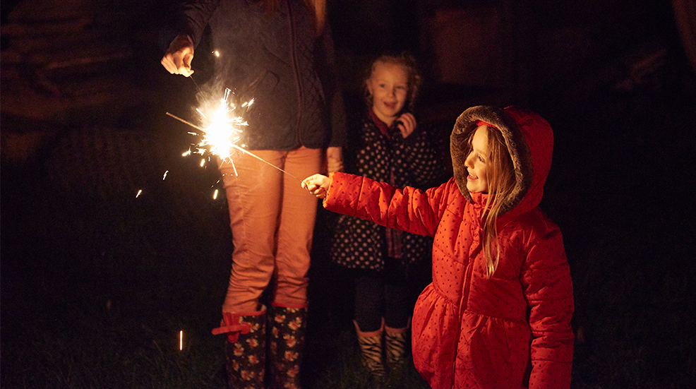 Side view of girl wearing red fur trim coat holding sparkler being ignited by mother